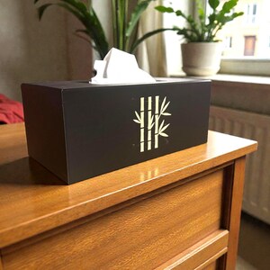 May include: A black tissue box featuring a gold bamboo design. White tissues protrude from the top. The box rests on a wooden dresser. Plants are visible in the background.