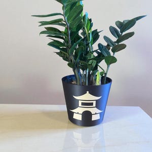 May include: A ZZ plant with dark green, glossy, oval-shaped leaves growing in a matte black cylindrical planter. The front of the planter features a white graphic illustration of a traditional pagoda structure with a tiered roof and arched entrance.