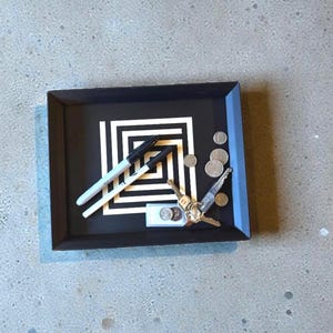 May include: A black rectangular tray with a geometric black and white design. Two white and black markers, coins, and keys are inside the tray. The tray sits on a gray surface.