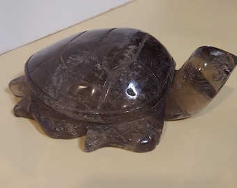 May include: A carved, dark gray crystal turtle figurine. The turtle has a detailed shell pattern and visible legs, head, and tail. The figurine is made of a translucent, smoky quartz-like material, and sits on a light beige surface.