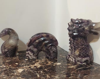 May include: Three carved amethyst sculptures. The first is a curved, abstract form. The second is a rounded, arch-shaped sculpture. The third is a dragon head with a detailed, textured body. All are shades of purple and white.