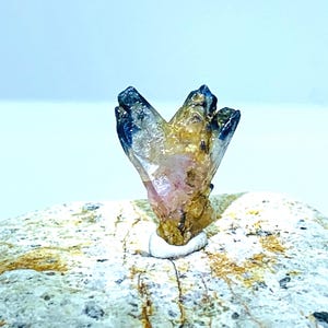 Peut inclure: Un amas de cristaux aux pointes bleu foncé et au corps translucide, rose doré. Les cristaux sont montés sur une roche claire et texturée. Le fond est bleu clair.