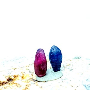 Peut inclure: Deux pierres précieuses brutes, l'une d'un magenta profond et l'autre d'un bleu vibrant, sont présentées sur une surface de pierre claire. Les gemmes ont une texture naturelle rugueuse et se dressent, mettant en valeur leurs formes et couleurs uniques.