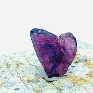 Può includere: Una gemma grezza a forma di cuore, di colore viola, con una consistenza ruvida. La gemma è posta su una superficie rocciosa chiara e strutturata, con uno sfondo bianco.