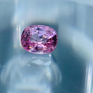 Puede incluir: Una gema rosa facetada de corte ovalado. La gema es de un tono rosa vibrante, con múltiples facetas que reflejan la luz. El fondo es azul claro, creando un contraste que resalta el color y el brillo de la gema.