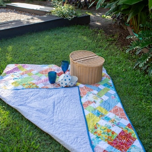 May include: A colorful picnic scene on a green lawn. A patchwork quilt with a blue border is spread out, with a wicker basket, two blue glasses, and a white bowl with blue polka dots. Lush greenery and a blue chair are in the background.