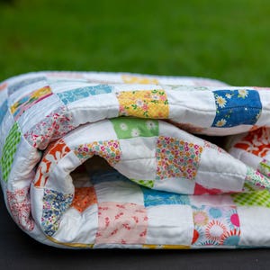 May include: Folded patchwork quilt with a white background and colorful square patterns. The squares feature floral and geometric designs in shades of blue, yellow, orange, and green. The quilt is folded over, showing the detailed stitching.