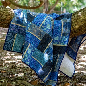 May include: A blue and teal quilt with various patterns, including swirls and geometric shapes, is draped over a tree branch. The quilt has a dark blue border and gold accents, creating a visually appealing textile.