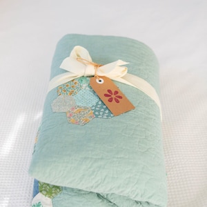 May include: A folded sage green quilt featuring a patchwork of small hexagonal fabric pieces with floral patterns. The quilt is tied with a cream-coloured ribbon and includes a brown paper tag with a red flower icon printed on the front.