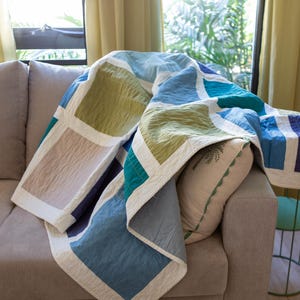 May include: A colourful quilted blanket with a geometric design in blues, greens, and beige, draped over a grey sofa. The blanket has a white border and is paired with a decorative pillow. The scene is lit by natural light.
