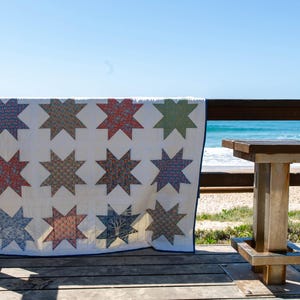 May include: A rectangular quilt featuring a white background with a grid of twelve eight-pointed stars. Each star is crafted from fabric with distinct patterns and colours, including shades of blue, red, green, and orange. The quilt has a dark blue border.
