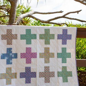 May include: A white quilt featuring a repeating grid of twelve cross-shaped patches. Each cross is made from fabric with distinct floral and geometric patterns in shades of pink, blue, green, and yellow. The quilt has a thin, patterned border.
