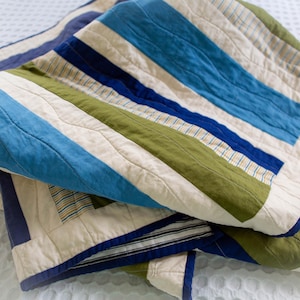 May include: A folded quilt with a patchwork design. The quilt features alternating stripes of blue, green, and cream-coloured fabric. The edges are trimmed with a dark blue border. The quilt is made of soft fabric and has a quilted pattern.