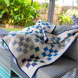May include: A handmade quilt with a blue and white checkered pattern, draped over a grey outdoor sofa. The quilt features squares of varying shades of blue and white, with a dark blue border. The sofa has striped cushions and is set against a backdrop of greenery.