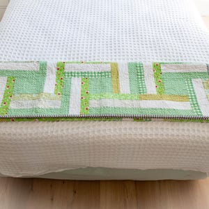 May include: A patchwork quilt in green, white, and yellow hues, featuring gingham and floral patterns. The quilt has a black and white striped border. It is displayed on a white bed. The quilt's design is reminiscent of a traditional craft.