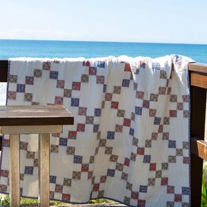 May include: A rectangular quilt featuring a white background with a repeating chain pattern composed of small squares in shades of blue, red, and green. The fabric has a visible stitched texture throughout, with a thin blue border along the edges.