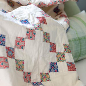 May include: A patchwork quilt featuring a repeating pattern of solid off-white squares alternating with smaller squares printed in floral motifs of red, blue, green, and pink. The textile is constructed with visible stitching lines between each square.