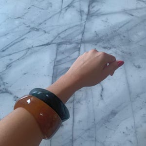 May include: Two smooth, wide bangle bracelets worn on the wrist. One bangle is a translucent, warm amber-orange color, while the other is a solid, opaque dark forest green. Both bracelets feature a rounded, polished surface and a thick, chunky design.