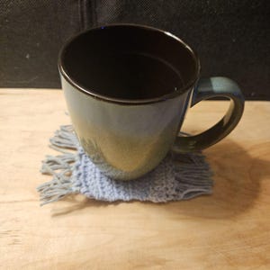 Puede incluir: Una taza de cerámica azul y marrón con interior oscuro se encuentra sobre un posavasos de ganchillo azul claro con flecos. La taza tiene un asa curva y una combinación de colores degradados, que va del azul al marrón.