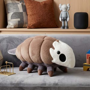 Puede incluir: Un gran peluche con forma de cochinilla, con un cuerpo marrón segmentado, patas grises y una cabeza blanca con ojos negros. El juguete está sobre un sofá gris, con cojines decorativos y figuritas en el fondo.