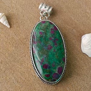 Puede incluir: Colgante ovalado con una piedra preciosa verde y roja engastada en un marco de metal plateado. El colgante tiene una fianza decorativa y se muestra sobre una superficie arenosa, con conchas marinas en el fondo.