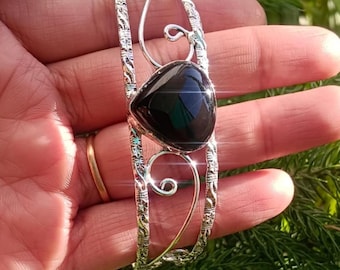 May include: A silver-toned cuff bracelet with an ornate design and a large, polished black gemstone at the center. The bracelet features decorative swirls and a textured pattern along the edges, creating an elegant accessory.