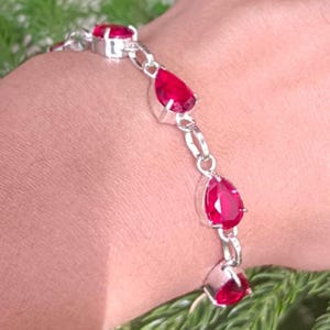 Puede incluir: Una pulsera de plata con piedras preciosas rojas vibrantes. La pulsera tiene un diseño de cadena con piedras redondas y en forma de lágrima alternadas. Las piedras están engastadas en biseles de plata.