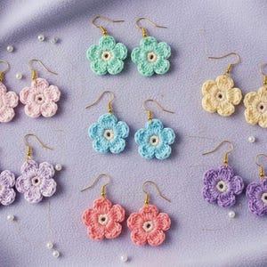 Puede incluir: Pendientes de flores de ganchillo hechos a mano en varios colores, incluyendo rosa, verde, azul, amarillo, morado y coral. Cada par tiene un gancho dorado. Los pendientes están colocados sobre un fondo de tela lavanda con perlas dispersas.
