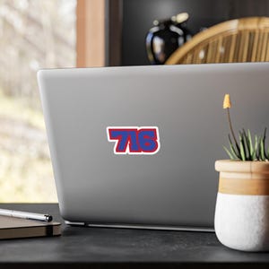 Op de afbeelding: Een sticker met het nummer "716" in rood en blauw, omlijnd met wit. De sticker is bevestigd op een grijze laptop. De laptop staat op een zwart oppervlak, met een kleine potplant en een notitieboek.