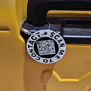 May include: A silver luggage tag with the text "CONTACT ME TO SCAN" around a QR code. The tag is attached to a yellow suitcase handle. The tag is round and has a silver wire loop.