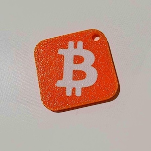 May include: Orange, square-shaped Bitcoin keychain with a white Bitcoin symbol. The keychain has a textured surface and a small hole at the top for attachment. The Bitcoin symbol is centered on the orange background.