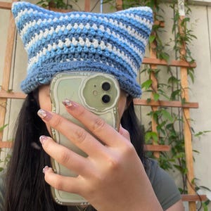 May include: A handmade crochet hat in shades of blue and white, featuring pointed cat ears. The hat is a bucket style, with a brim and a striped pattern. The hat is worn by a person.