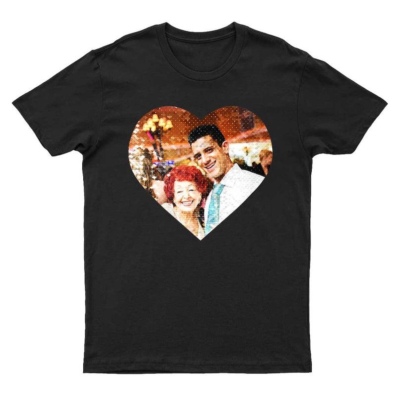 May include: Black t-shirt with a heart-shaped image of a man and a woman smiling. The photograph appears to be taken at an event. The background is blurred. The t-shirt is made of a soft material. The image is in the center of the t-shirt.