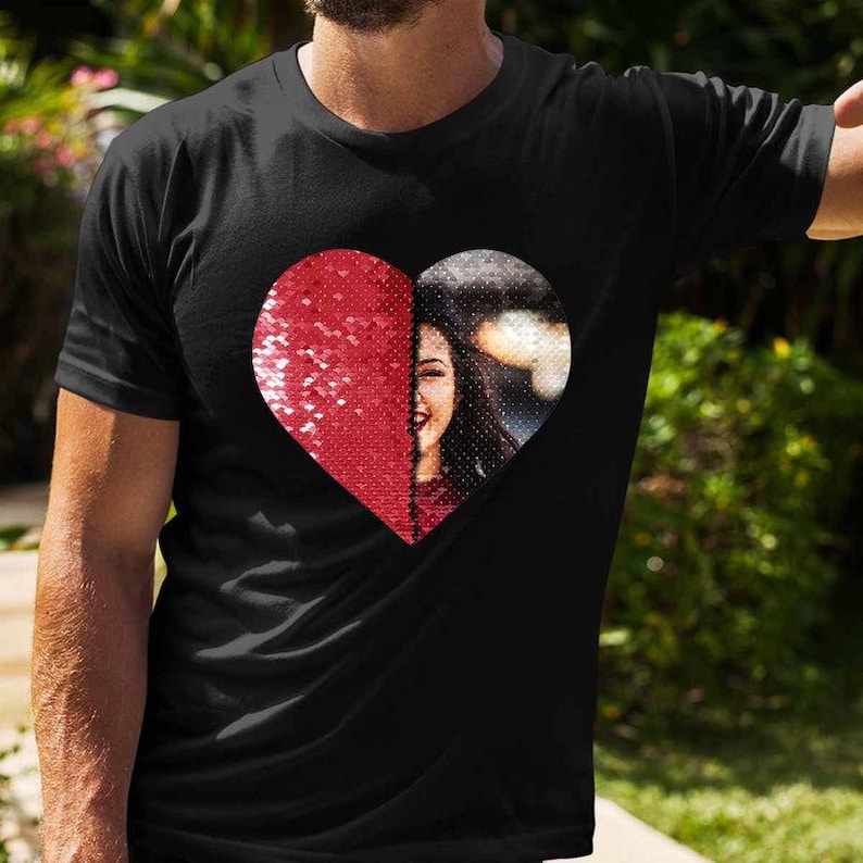 Puede incluir: Camiseta negra con dise&ntilde;o de coraz&oacute;n. La mitad del coraz&oacute;n est&aacute; cubierta de lentejuelas rojas, la otra mitad muestra a una mujer sonriendo. La camiseta es de estilo casual.