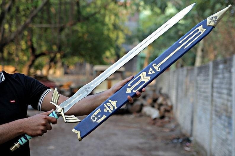 May include: A silver sword with a detailed hilt and a blue scabbard with gold accents. The sword is held next to its scabbard, showcasing the design. The scabbard has a gold emblem on the end.