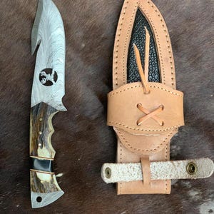 May include: A hunting knife with a patterned steel blade and a deer antler handle. The knife has a hook at the tip and a leather sheath with a belt loop. The sheath is tan and has a black interior.