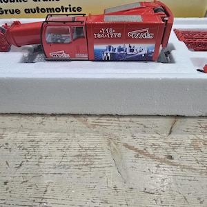 Puede incluir: Modelo rojo de grúa móvil con el texto "CRANES" y un número de teléfono en el lateral. El modelo está en un inserto de espuma blanca, con la caja parcialmente visible en el fondo. La grúa tiene un diseño detallado.