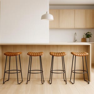 Puede incluir: Cuatro taburetes de bar de cuero marrón con patas de metal negro están colocados frente a una encimera de madera clara. Los taburetes tienen un asiento curvo y acolchado con un diseño acanalado. La cocina tiene armarios de madera clara y una pared blanca.