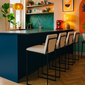 Peut inclure: Une cuisine moderne avec un îlot bleu marine et quatre tabourets de bar crème avec des cadres en métal noir. La cuisine comprend une crédence en carrelage turquoise, des étagères en bois et une suspension jaune. Le sol est en chevrons clairs.