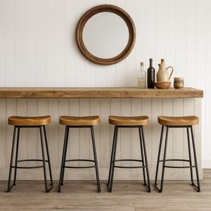 May include: Four wooden bar stools with black metal legs are arranged in front of a wooden bar. A round wooden mirror hangs on the white wall above the bar. The bar is decorated with bottles, a pitcher, and a small bowl.