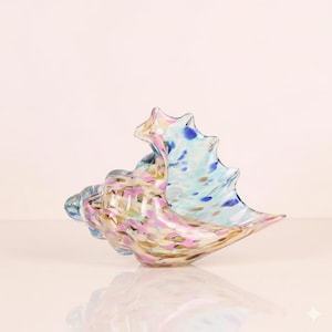May include: A colorful glass seashell sculpture with a speckled design. The sculpture features shades of pink, blue, and gold. The seashell is a decorative item, perfect for coastal or beach-themed decor.