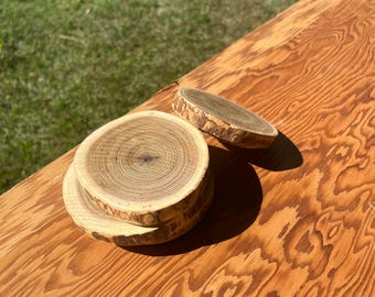 May include: A stack of three wooden coasters with natural bark edges and visible growth rings. A fourth coaster rests nearby. The coasters are made of light brown wood and sit on a wooden surface.