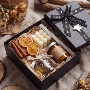 Op de afbeelding: Zwarte geschenkdoos met zwart lint met de tekst "BESTLOVE" en een gouden ster. Bevat een bubbelkaars, een kaars in een pot, een parfumfles, kaneelstokjes en gedroogde sinaasappelschijfjes.