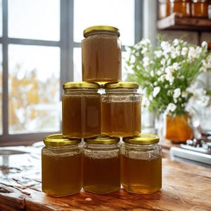 Puede incluir: Seis tarros de vidrio llenos de miel dorada, cada uno con una tapa dorada, apilados sobre una superficie de madera. La miel es de un rico color ámbar. El fondo incluye una ventana y un jarrón con flores blancas.