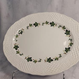 Puede incluir: Plato ovalado blanco para servir con un borde texturizado estilo mimbre. El centro presenta un anillo de diseños de hojas verdes y marrones. El plato está sostenido por un soporte metálico.