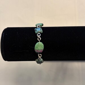May include: A handmade bracelet featuring natural gemstones. The bracelet has three green and blue stones connected by silver-colored links. The stones have a mix of colors, including green, blue, and red.