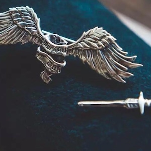 May include: Silver-coloured metal brooch and dagger set. The brooch features a winged skull design with a banner. The dagger has a cross-shaped handle. The set is displayed on a dark blue surface.