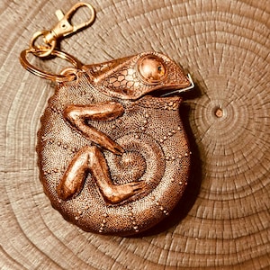 May include: A copper-colored chameleon keychain with a gold-colored clasp and key ring. The reptile has detailed scales and a curled tail. The keychain is made of metal and is a decorative accessory.