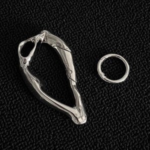 Puede incluir: Un mosquetón plateado para llavero y un pequeño anillo redondo, ambos de metal. El clip tiene un diseño orgánico y fluido con una abertura con bisagras. El anillo tiene un diseño simple y circular. Ambos artículos se muestran sobre una superficie negra texturizada.