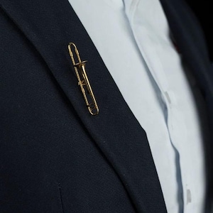 Pode incluir: Um alfinete de lapela dourado em forma de trombone está preso a um blazer azul marinho. O alfinete é uma réplica em miniatura do instrumento musical, com detalhes precisos. Uma camisa branca é visível por baixo.